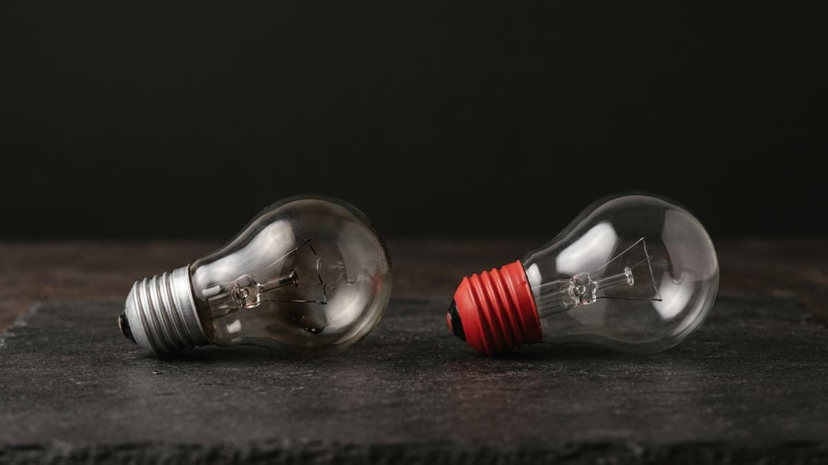 A burnt-out light bulb lying on a dark slate surface beside a new bulb with a red base, ready to take its place.